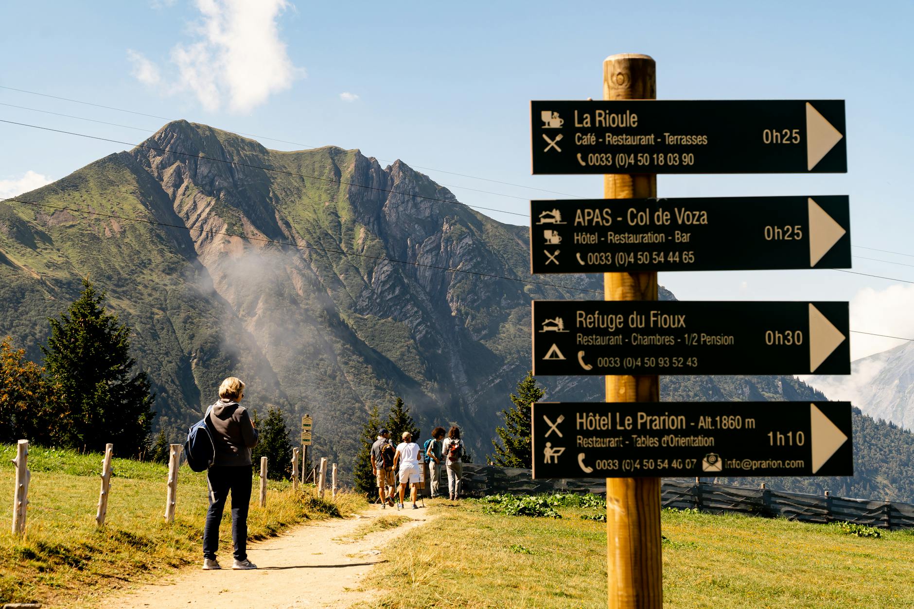 Une famille en randonnée sur un sentier de montagne ensoleillé en France, avec enfants et sac à dos, paysage verdoyant en arrière-plan